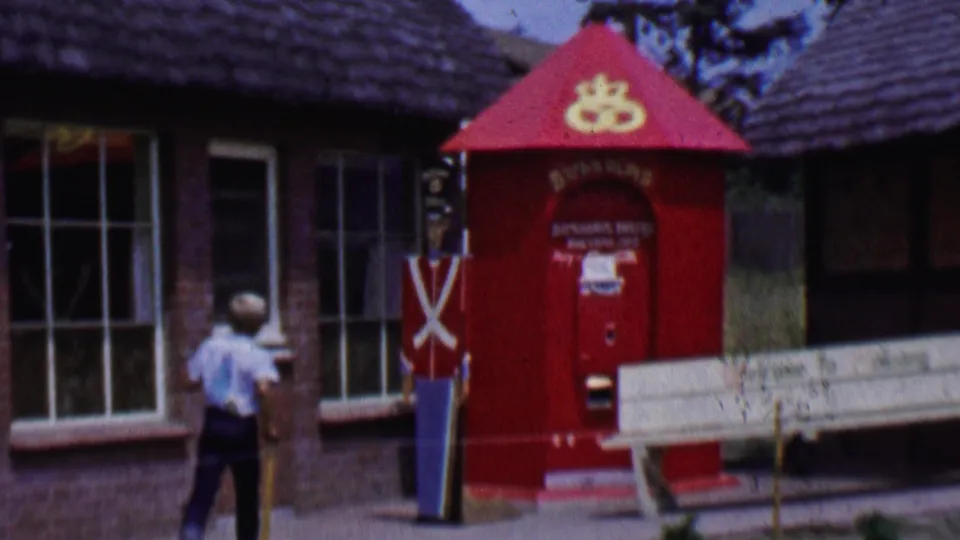 1955 Solvang street scene with the walker beside a red kiosk, toy soldier figure, white bench, brick storefront, and thatched roofline.