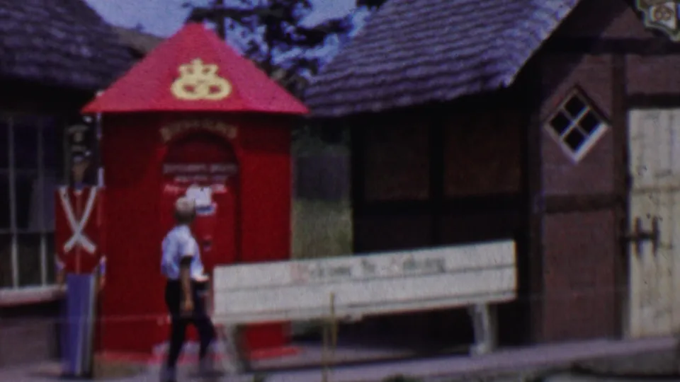 1955 Solvang footage showing the walker near the red kiosk and white bench, with a shop window and timbered building facade behind him.