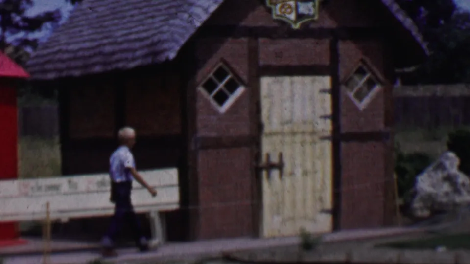 1955 Solvang street view with the walker passing a timbered facade, pale double doors, small diamond windows, a thatched roof, and a white bench.