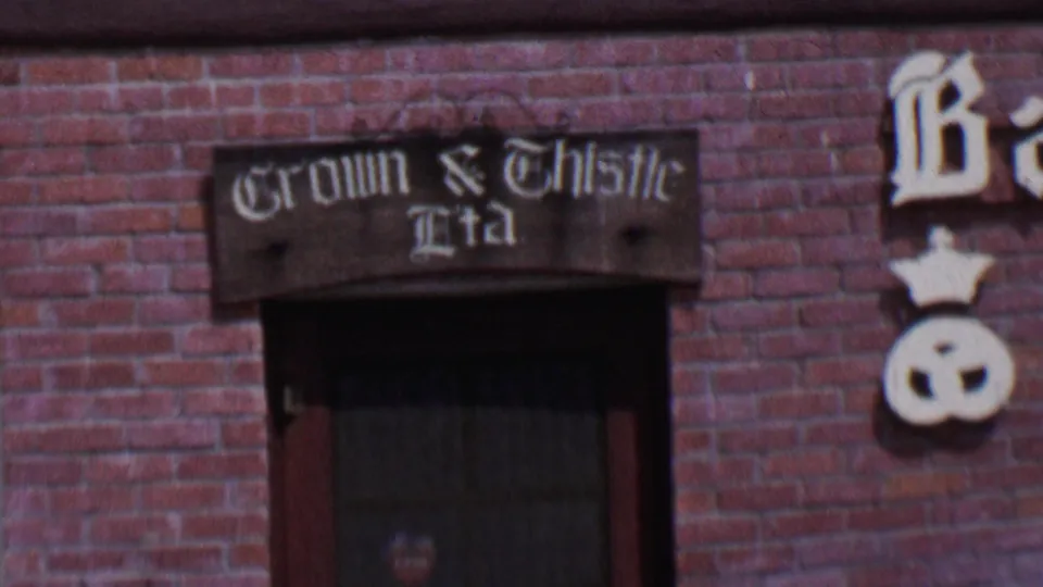 1955 Solvang, California storefront detail with Crown & Thistle Ltd lettering, a dark window, and a white crown-and-pretzel symbol on the brick wall.