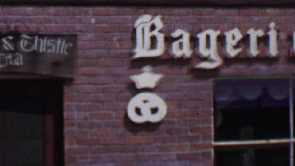 1955 Solvang, California storefront detail showing partial Bageri lettering, Crown & Thistle signage, white pretzel emblems, and a window set into the brick wall.
