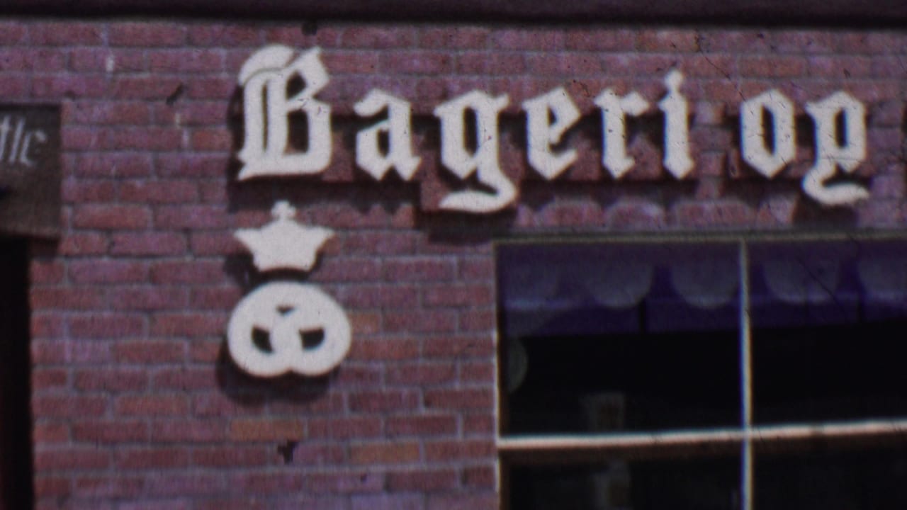 1955 Solvang, California 8mm frame showing the left portion of a Danish-style bakery facade with Bageri og lettering, brickwork, windows, and a crown-topped pretzel sign.