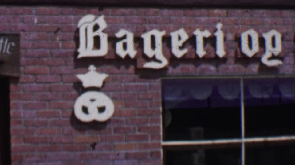 1955 Solvang, California 8mm frame showing the left portion of a Danish-style bakery facade with Bageri og lettering, brickwork, windows, and a crown-topped pretzel sign.