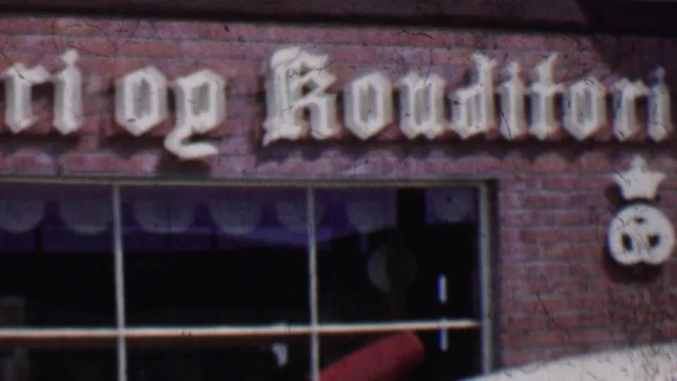 1955 Solvang, California 8mm frame showing the lower pan across the bakery facade, with partial Konditori lettering, brickwork, window glass, and a crown-topped pretzel emblem.