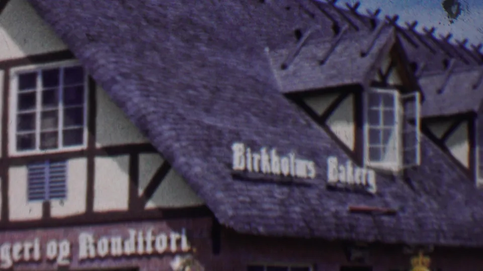 1955 Solvang, California 8mm frame showing the bakery roofline with timber-framed architectural details, small windows, and the Birkholms Bakery sign on the sloped roof.