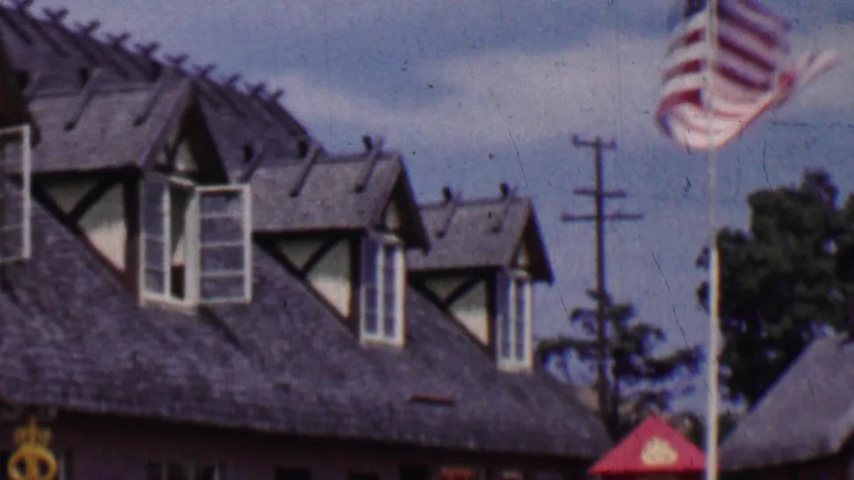 1955 Solvang, California exterior frame showing the bakery's steep roof and dormer windows with a U.S. flag, utility poles, and trees visible at the right edge.