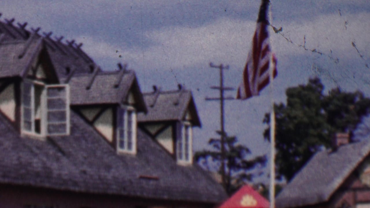1955 Solvang, California home-movie frame showing half-timbered rooflines, American flags on tall poles, trees, utility poles, and cloudy sky.