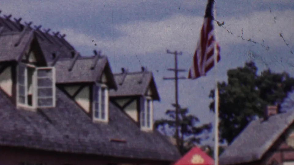 1955 Solvang, California home-movie frame showing half-timbered rooflines, American flags on tall poles, trees, utility poles, and cloudy sky.
