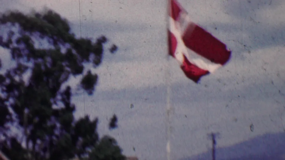 1955 Solvang, California home-movie frame showing a Danish flag on a tall pole with trees, cloudy sky, and distant hills behind it.