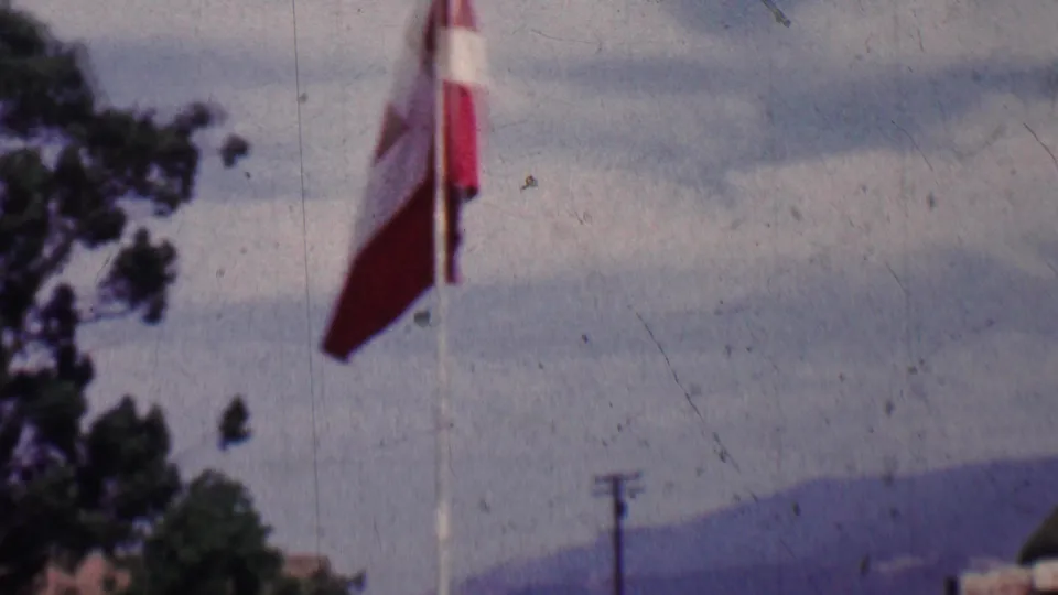 1955 Solvang, California frame showing a red and white flag on a tall pole against cloudy sky, distant hills, trees, and utility poles.