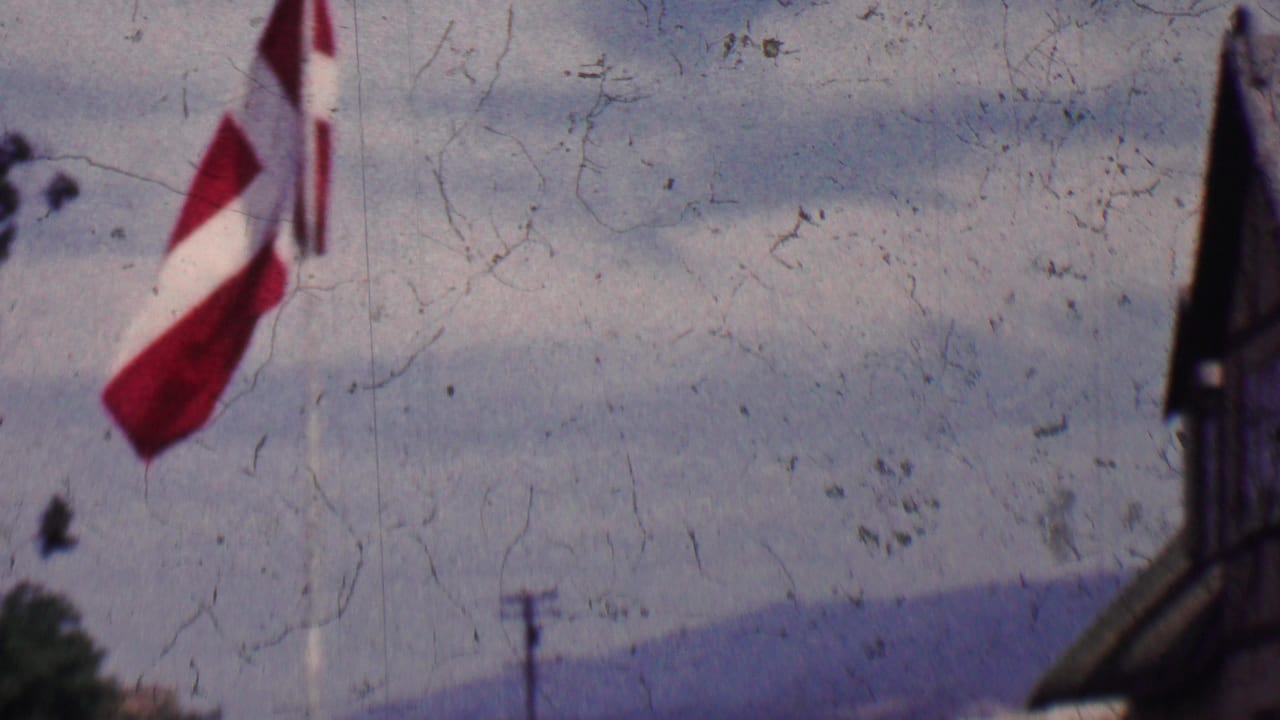 A red-and-white Danish flag flutters at the edge of a 1955 Solvang, California home-movie view with half-timbered architecture, distant hills, and cloudy sky.