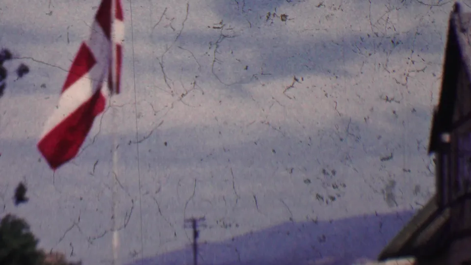 A red-and-white Danish flag flutters at the edge of a 1955 Solvang, California home-movie view with half-timbered architecture, distant hills, and cloudy sky.