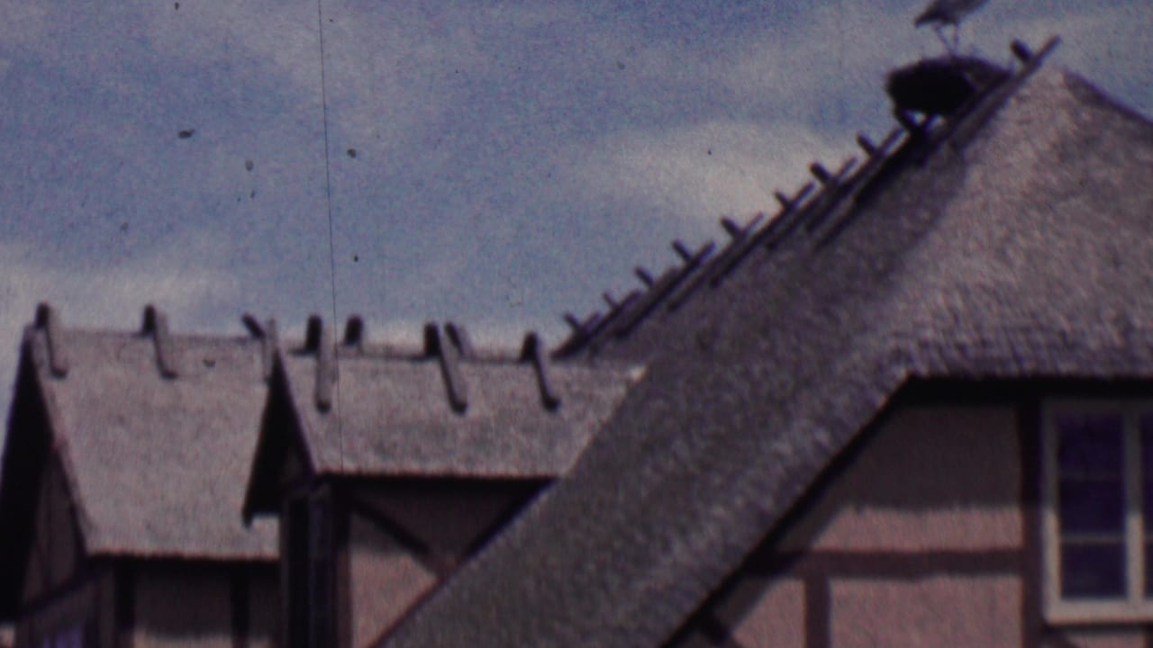 1955 Solvang, California home-movie frame showing steep thatched roofs, dormer windows, and timber-frame facade details from a visitor's upward street view.