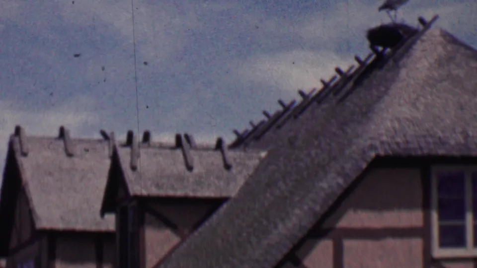 1955 Solvang, California home-movie frame showing steep thatched roofs, dormer windows, and timber-frame facade details from a visitor's upward street view.