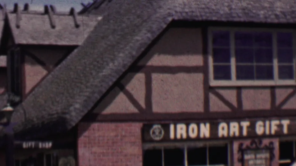 1955 Solvang, California home-movie frame looking toward a brick and timber-frame gift shop facade with thatched roof slopes and a partially visible IRON ART GIFT sign.