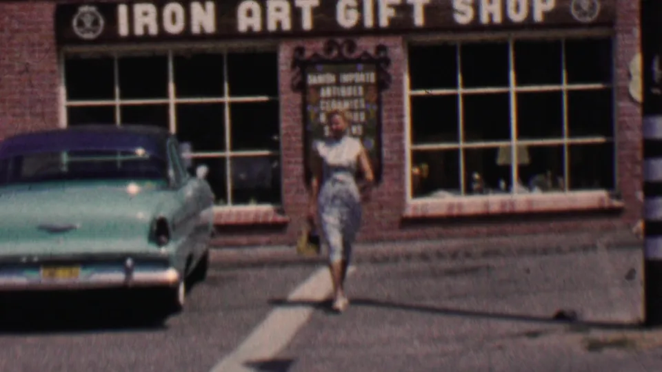 A woman in a light patterned dress walks in front of the Iron Art Gift Shop in Solvang, California, with a turquoise 1950s car parked beside the storefront.