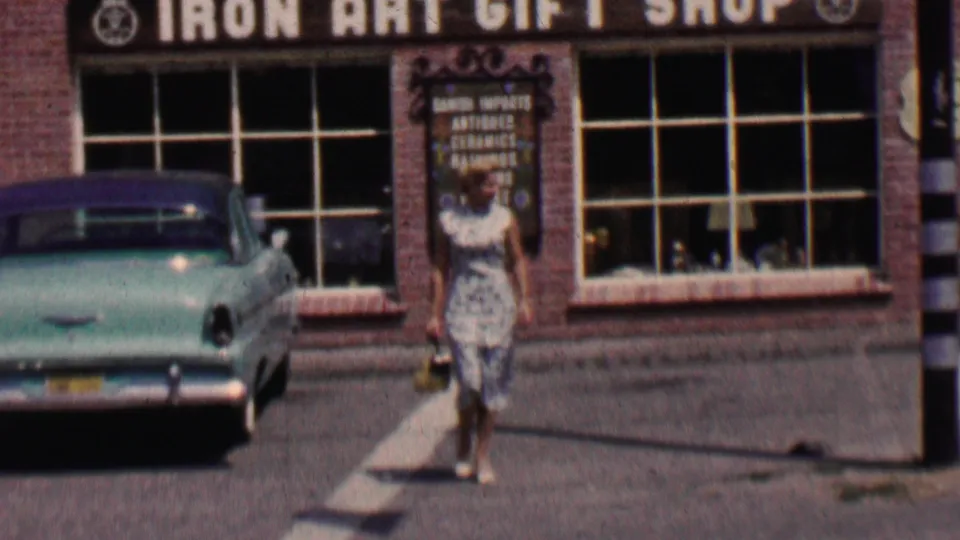 The woman continues walking past the Solvang Iron Art Gift Shop, carrying a purse near the display windows and parked turquoise mid-century car.