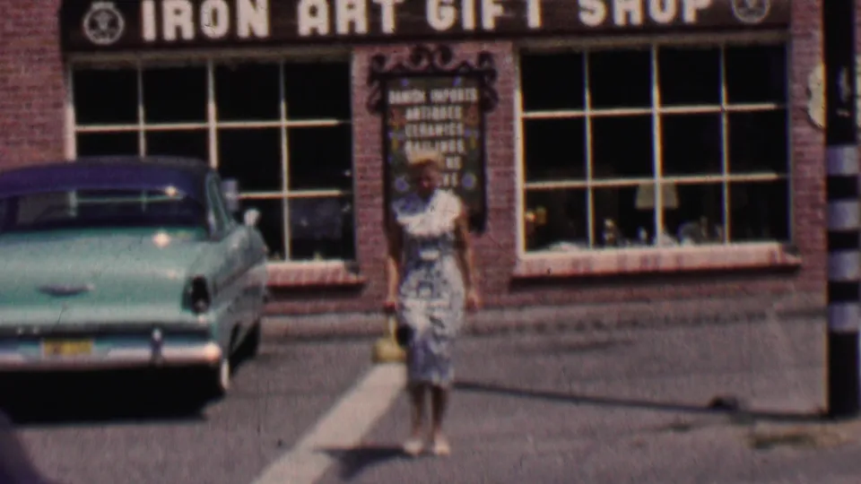 In Solvang, California, a woman in a patterned dress walks toward the camera outside the brick Iron Art Gift Shop while a turquoise 1950s sedan is parked along the street.