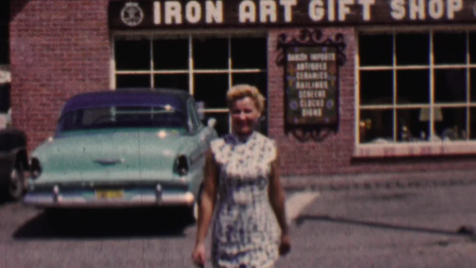 The woman walks closer to the camera in the Solvang street scene, with the Iron Art Gift Shop sign, brick wall, windows, and turquoise sedan visible behind her.