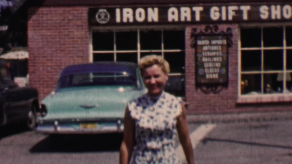 The woman fills more of the frame as she walks in front of the parked turquoise car, with the Iron Art Gift Shop facade and Danish imports sign partly visible behind her.