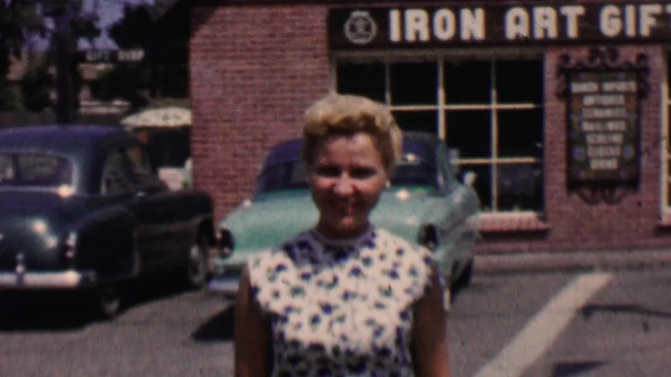 At the end of the clip, the woman stands or walks very close to the camera while the Solvang gift shop signage, brick storefront, turquoise sedan, and another car remain visible in the background.