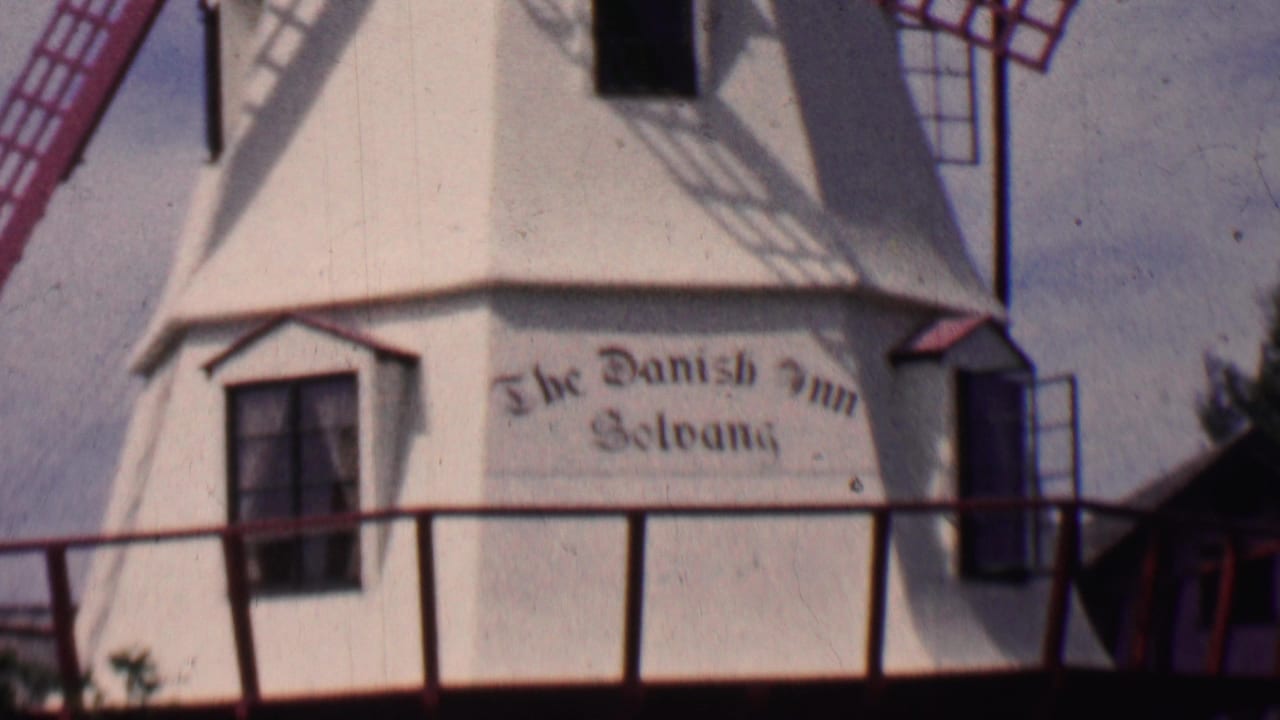 1955 Solvang, California home-movie frame showing a close view of a white Danish-style windmill with red lattice blades and the painted The Danish Inn Solvang sign.