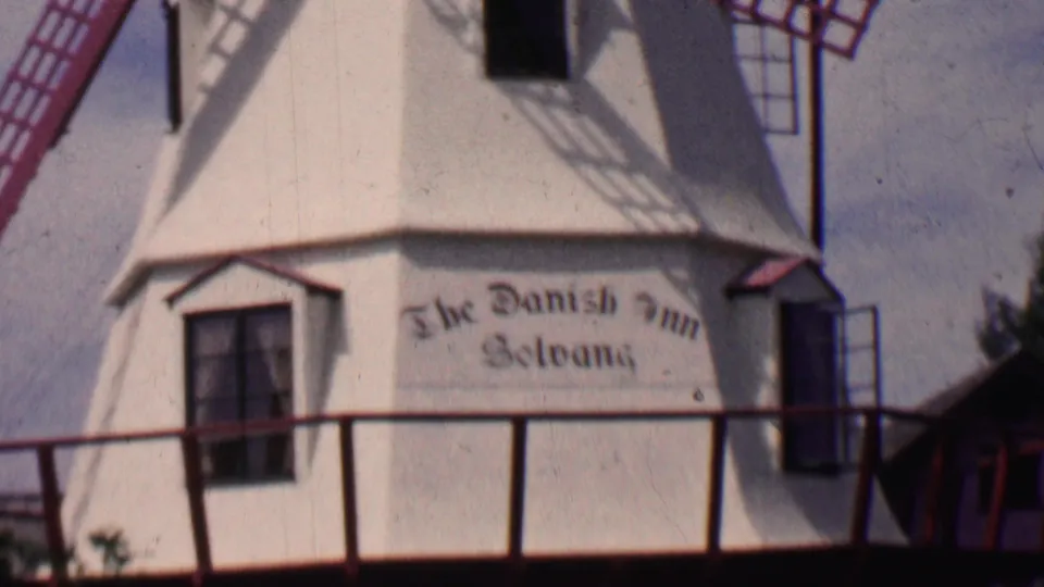 1955 Solvang, California home-movie frame showing a close view of a white Danish-style windmill with red lattice blades and the painted The Danish Inn Solvang sign.