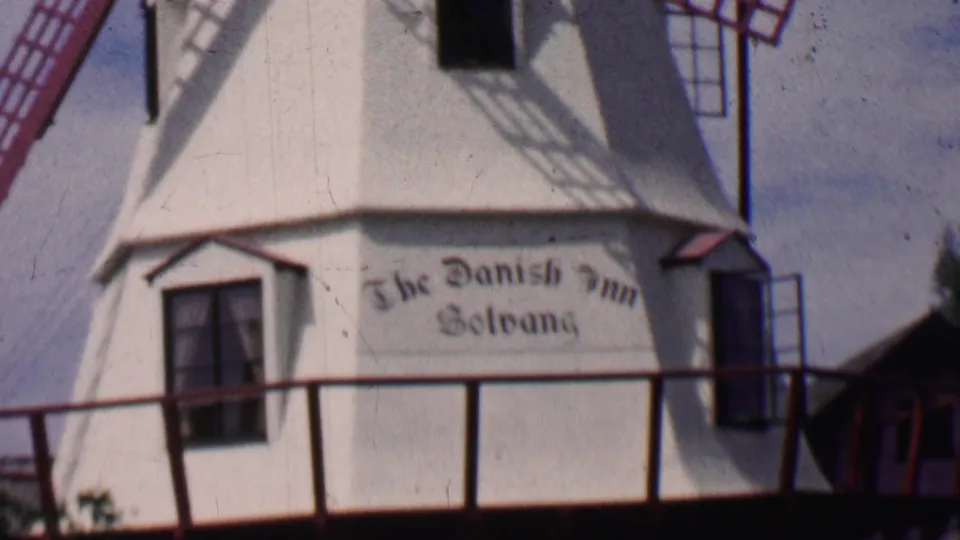 1955 8mm frame from Solvang showing the windmill facade cropped tightly around the window, red railing, and painted The Danish Inn Solvang sign.