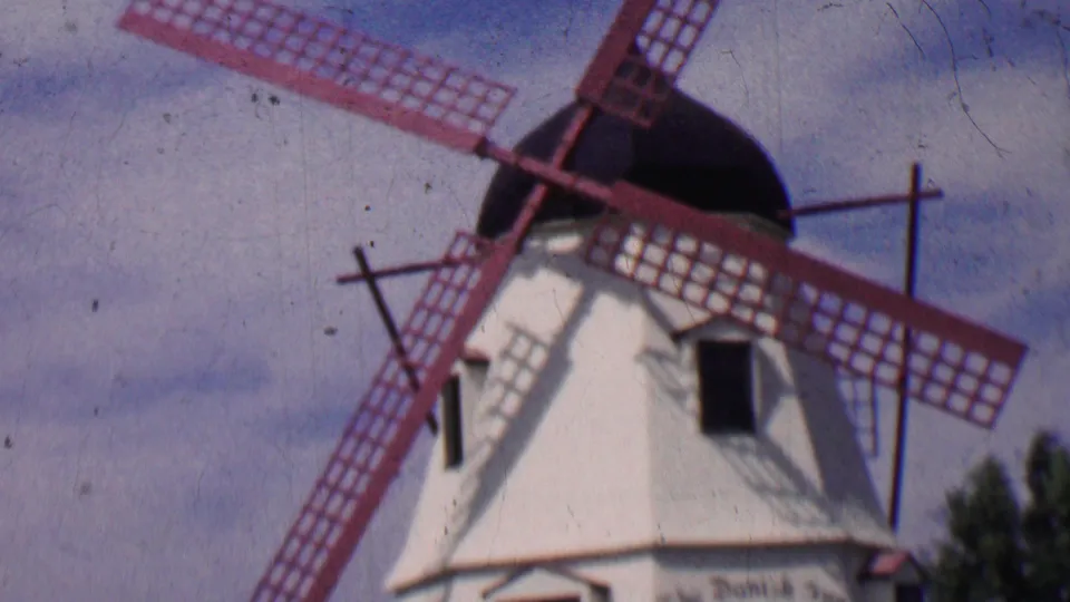 1955 Solvang frame showing the upper portion of the Danish-style windmill, red lattice blades, dark cap, white tower, and cloudy sky.