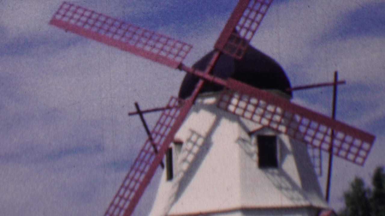 1955 Solvang home-movie frame showing a white Danish-style windmill with red lattice blades seen from a low angle against blue sky and clouds.