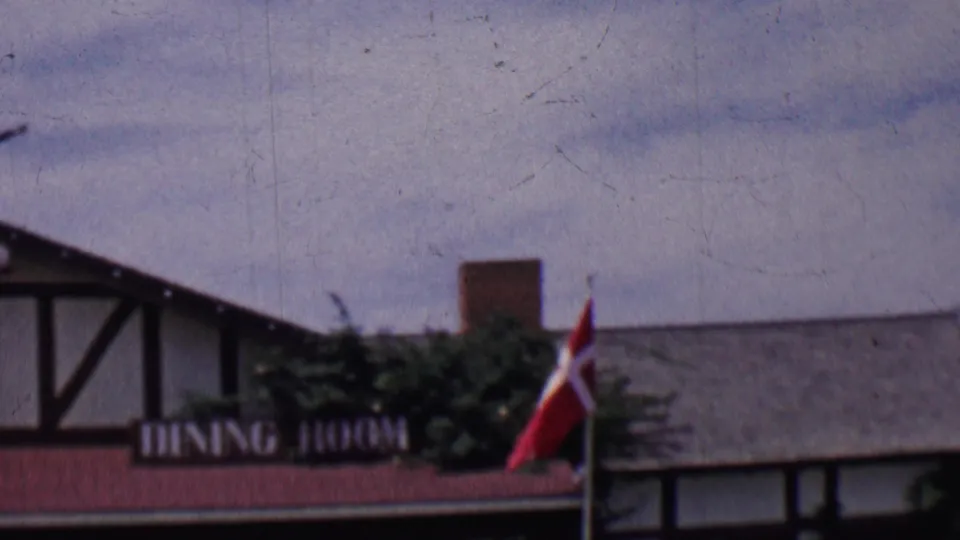 1955 Solvang, California 8mm frame showing a brown-and-white half-timbered dining room facade, red roofline, shrubs, chimney, and Danish flag.