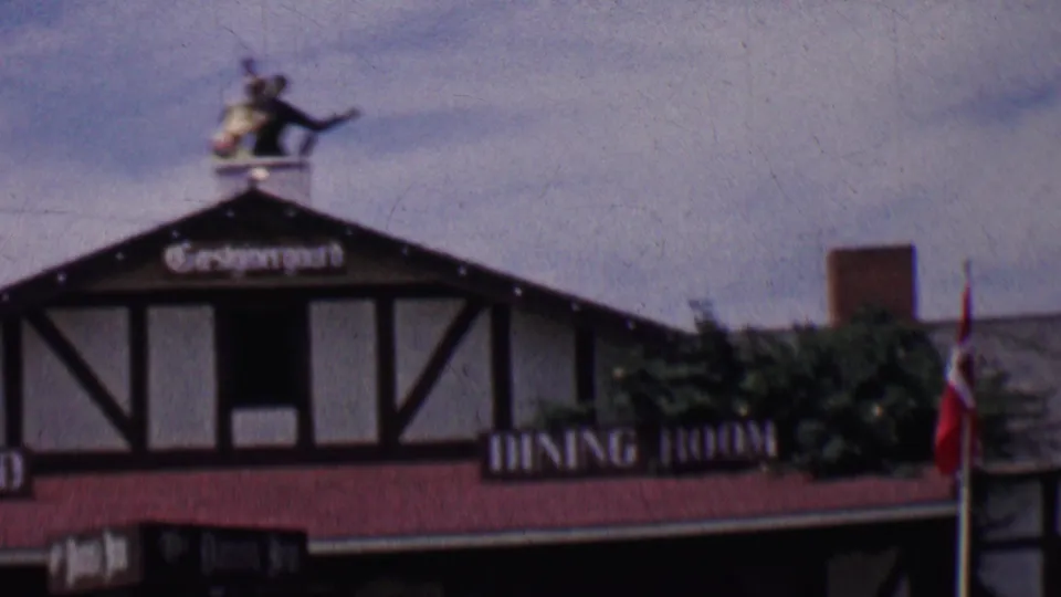1955 Solvang, California frame showing a half-timbered dining room building with a small rooftop figure, red trim, shrubs, and a Danish flag at the edge.