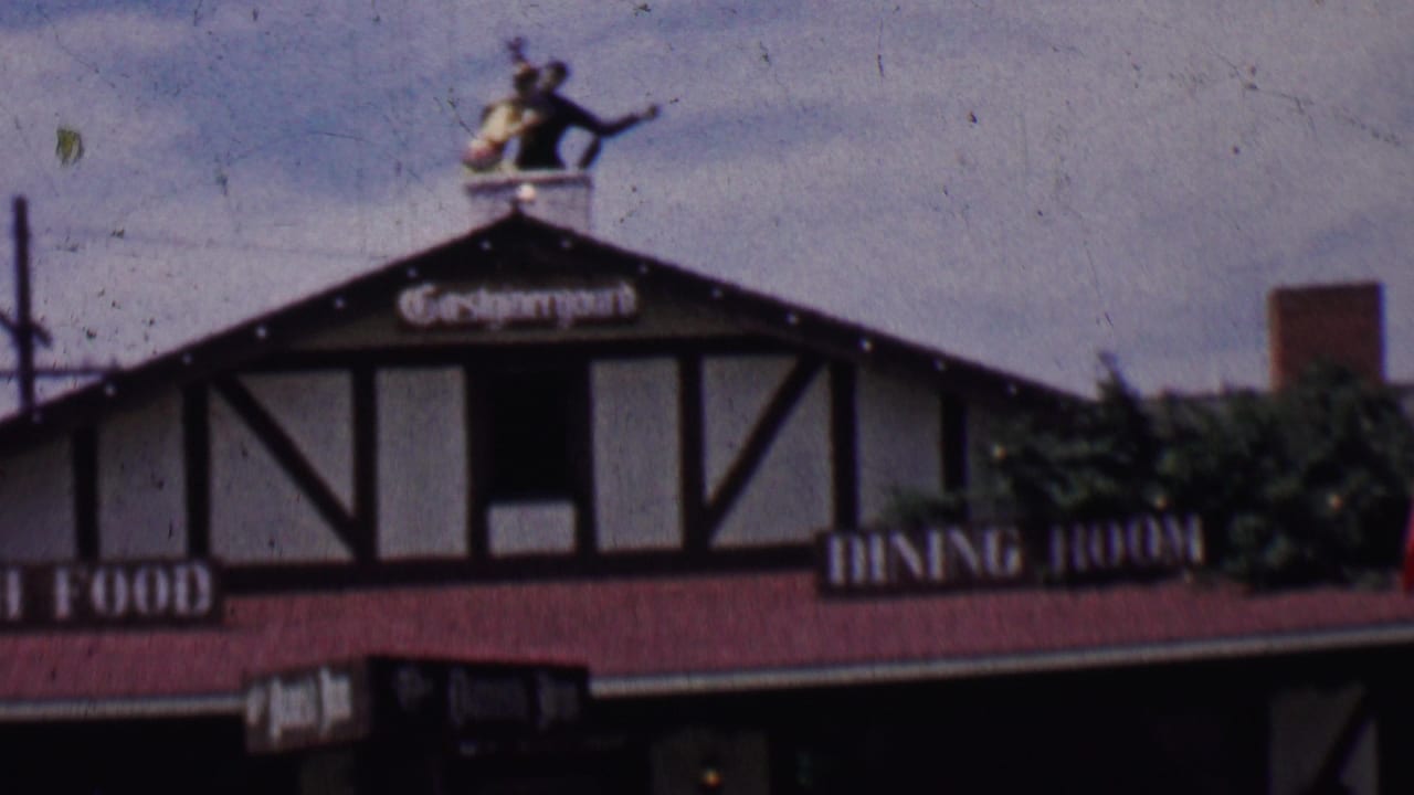 1955 Solvang, California home-movie frame showing a Danish-style restaurant facade with red roof, painted signage, rooftop figure, and utility lines.