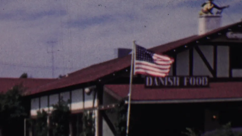 1955 Solvang, California home-movie frame showing an American flag in front of a Danish-style restaurant exterior with red roof and Danish Food sign.