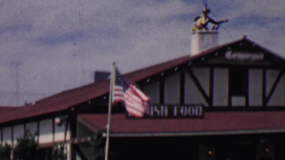 1955 Solvang, California frame showing the American flag, red-roofed half-timbered restaurant facade, Danish Food lettering, and rooftop figure.
