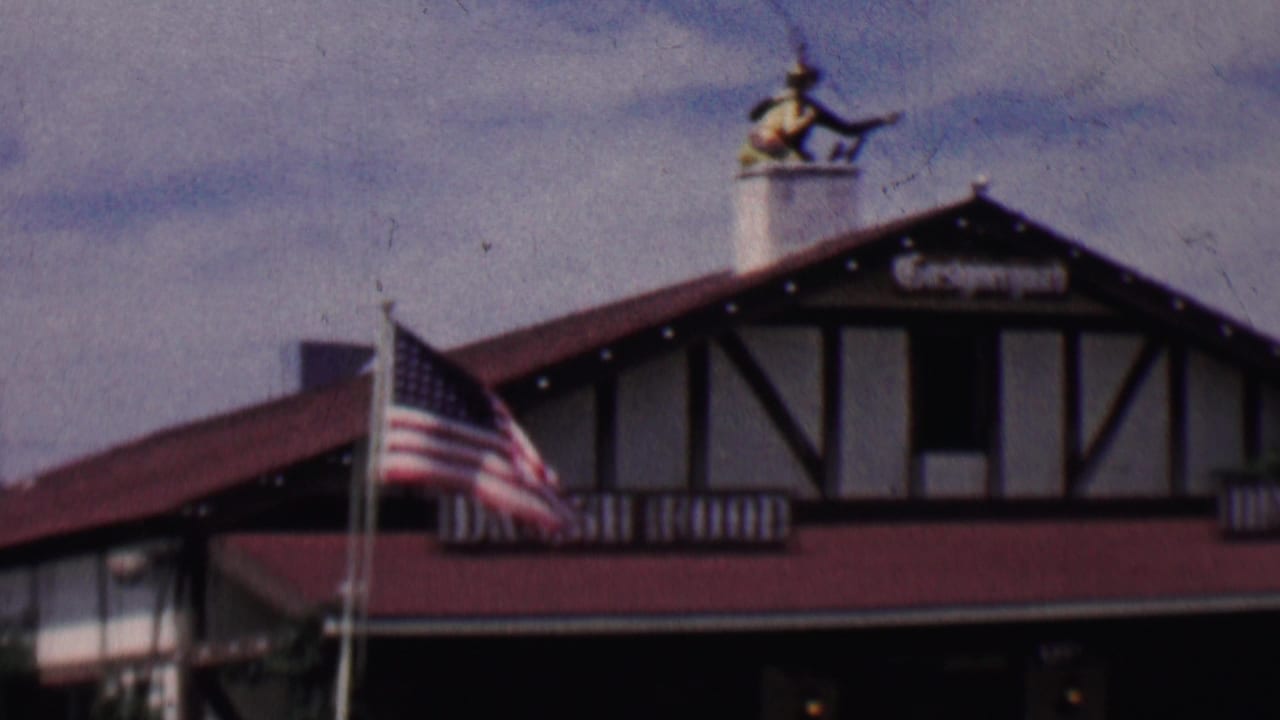 1955 Solvang, California home-movie frame showing an American flag before half-timbered Danish-style restaurant buildings with red roofs and visible Danish Food signage.