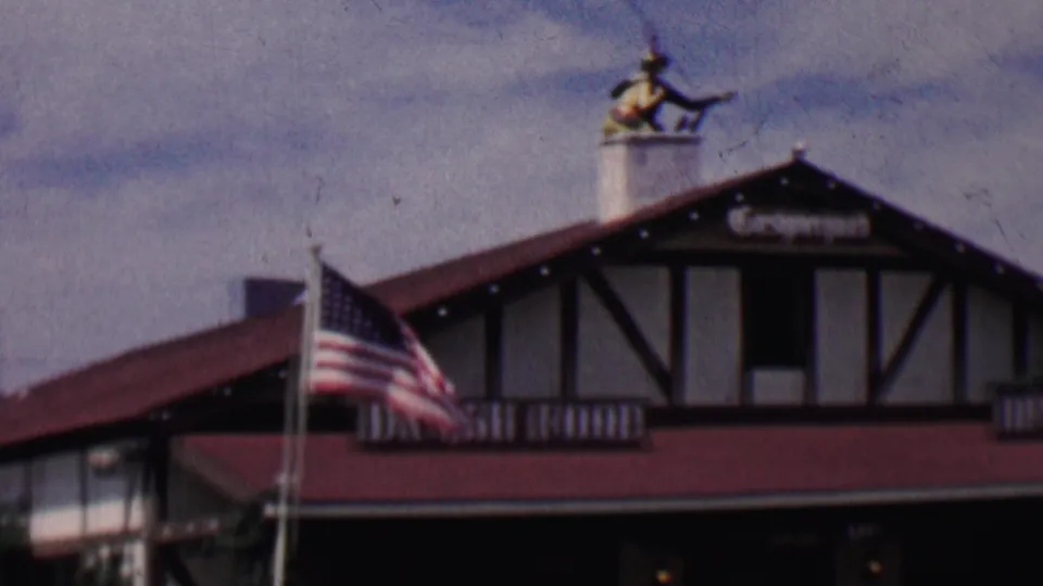 1955 Solvang, California home-movie frame showing an American flag before half-timbered Danish-style restaurant buildings with red roofs and visible Danish Food signage.