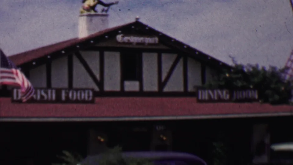 1955 Solvang, California frame showing an American flag waving before a Danish-themed restaurant facade with half-timbering, red roofing, and dining signage.