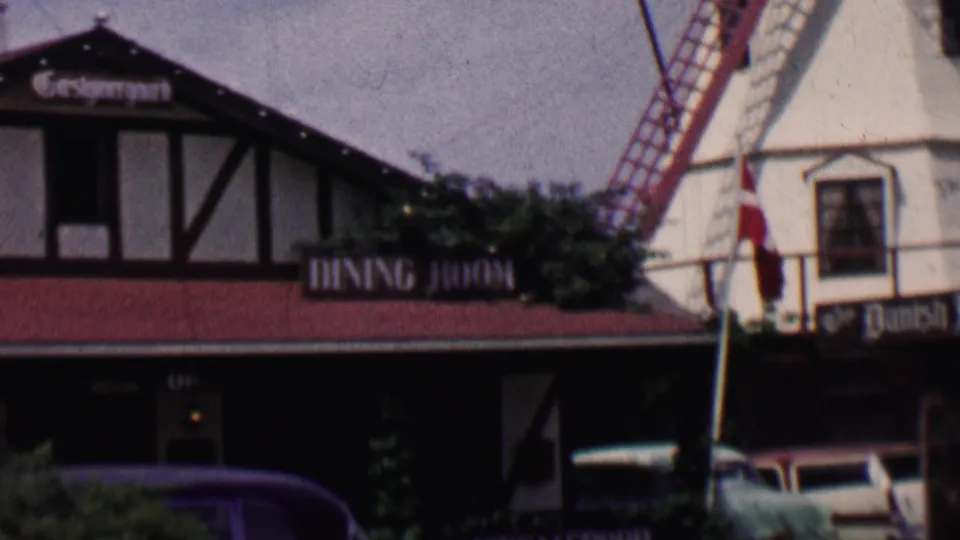 1955 Solvang, California frame showing the camera pan reaching a white windmill structure beside Danish-style restaurant buildings, flags, shrubs, and parked mid-century cars.
