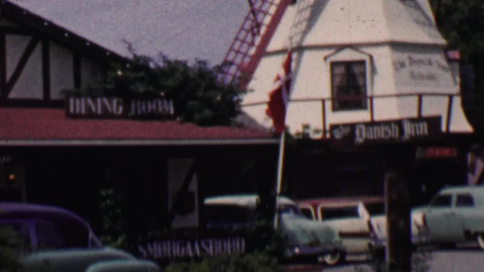 1955 Solvang, California frame showing a white windmill with red structural elements, Danish-themed dining signs, flags, and several parked 1950s automobiles in front.