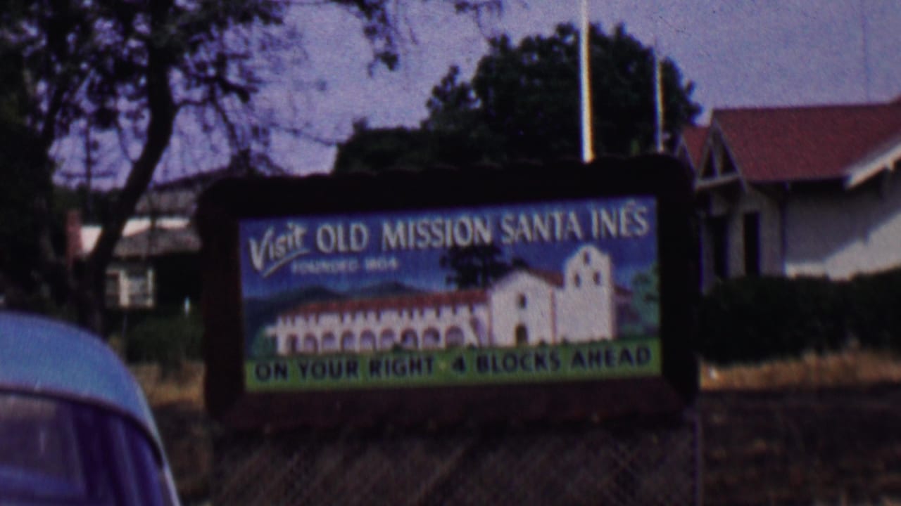 Large roadside sign inviting visitors to Old Mission Santa Ines with a building behind it and part of a car in the foreground.