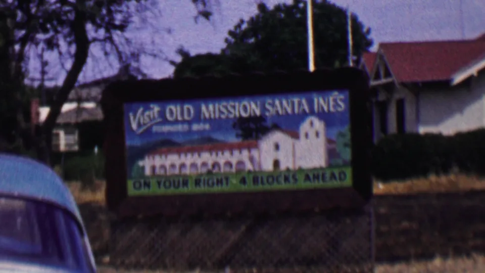 A 1950s Solvang visitor sign for Old Mission Santa Ines stands beside the road, with trees, dry grass, a nearby building, and a partial blue car at frame edge.