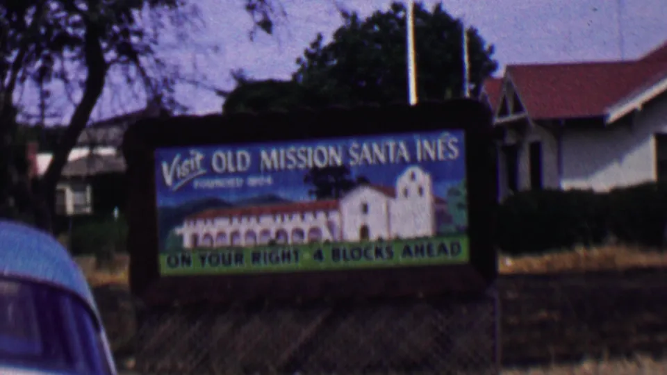 Solvang, California 1950s frame showing the Old Mission Santa Ines sign and illustrated mission facade, with trees, dry roadside grass, and neighboring gabled architecture.