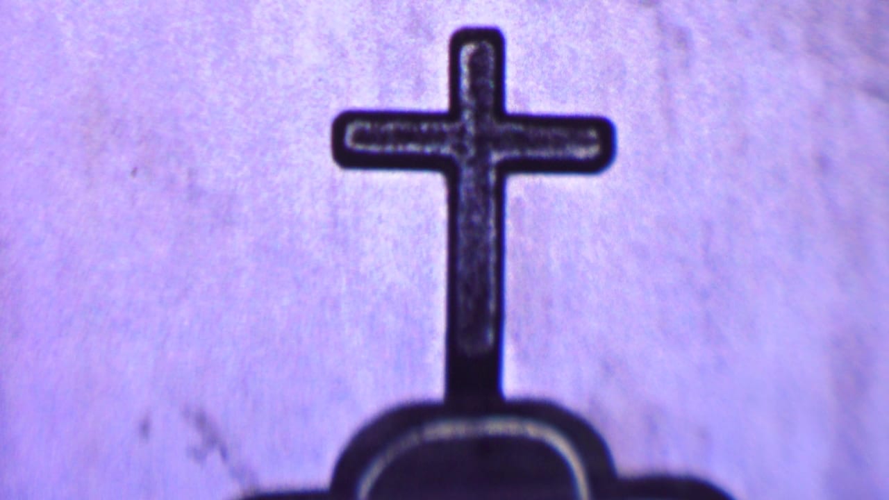 Close 1955 Solvang 8mm frame showing a dark cross rising from the top of a mission-related grave marker against a purple-tinted surface.