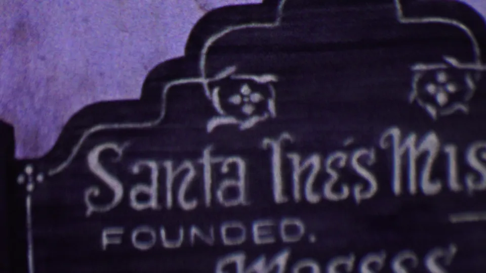 Close 1955 Solvang 8mm frame showing the Santa Ines Mission name, the word Founded, and decorative carving on a dark religious marker.