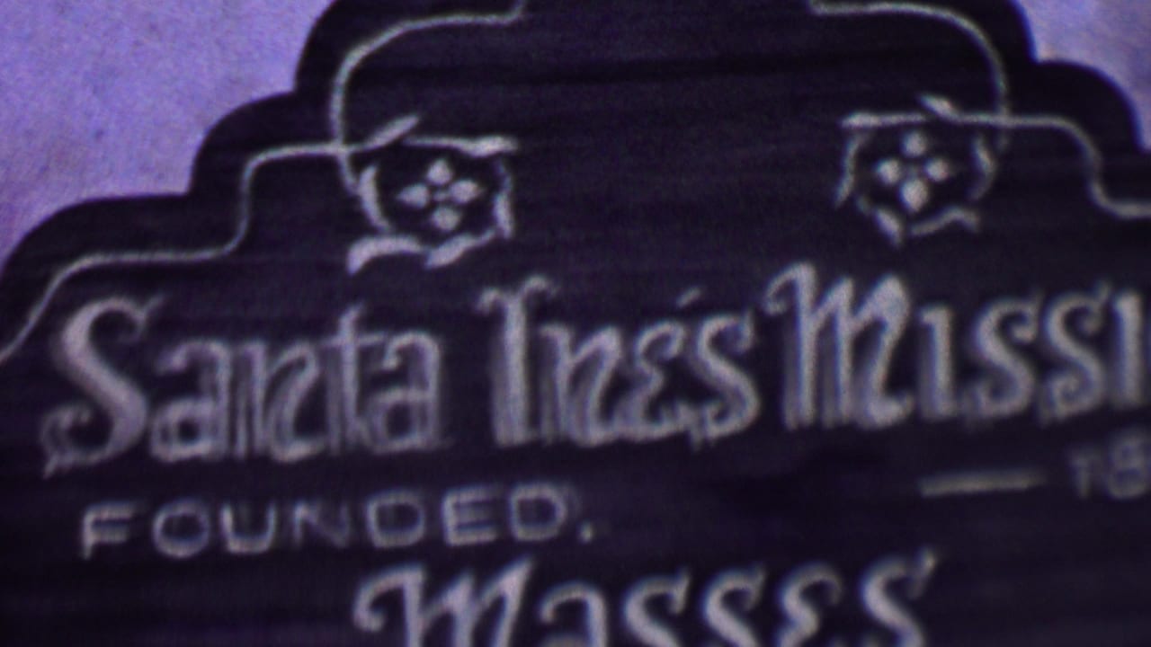 Soft 1955 8mm close-up of the Santa Inés Mission sign in Solvang, showing ornate white lettering, decorative floral details, and part of the founded text.