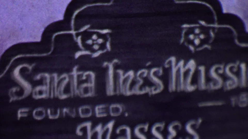 Soft 1955 8mm close-up of the Santa Inés Mission sign in Solvang, showing ornate white lettering, decorative floral details, and part of the founded text.