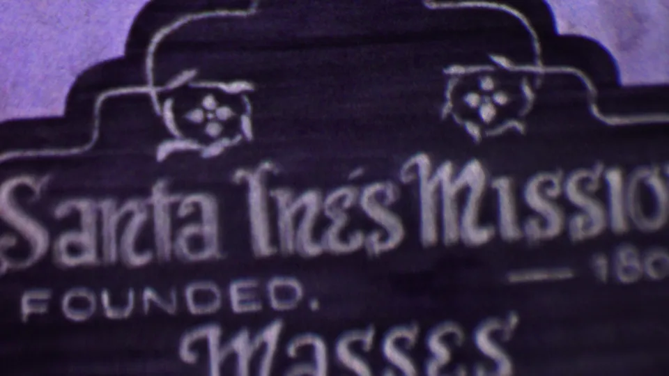 Close 8mm frame of the Santa Inés Mission sign, with the mission name, decorative border shape, and partial founding-year text visible in soft focus.