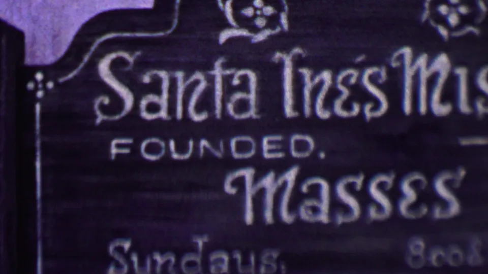 1955 Solvang 8mm frame showing the Santa Inés Mission sign with founded wording, large masses text, and partial Sunday schedule information.