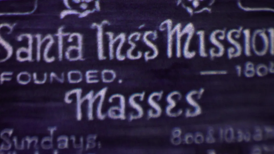 Final 8mm frame on the Santa Inés Mission sign in Solvang, showing ornate lettering, founded 1804 text, masses wording, and partial Sunday and weekday schedule lines.
